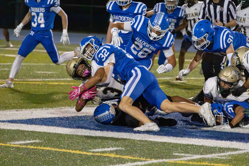 A football player in a blue uniform is tackling an opponent wearing a gold helmet during a game on a field. Other players are visible in the background.
