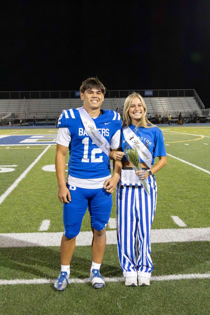 A male football player in a blue jersey with the number 12 stands next to a female wearing blue and white striped pants, holding a bouquet. Both are smiling on a football field at night.