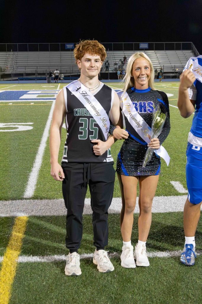 Two high school students posing on a football field during homecoming festivities. The male student is wearing a black jersey with the number 35 and a 'Homecoming Rep' sash, while the female student is in a sparkly blue cheerleading outfit with a 'Homecoming' sash, holding a flower bouquet.