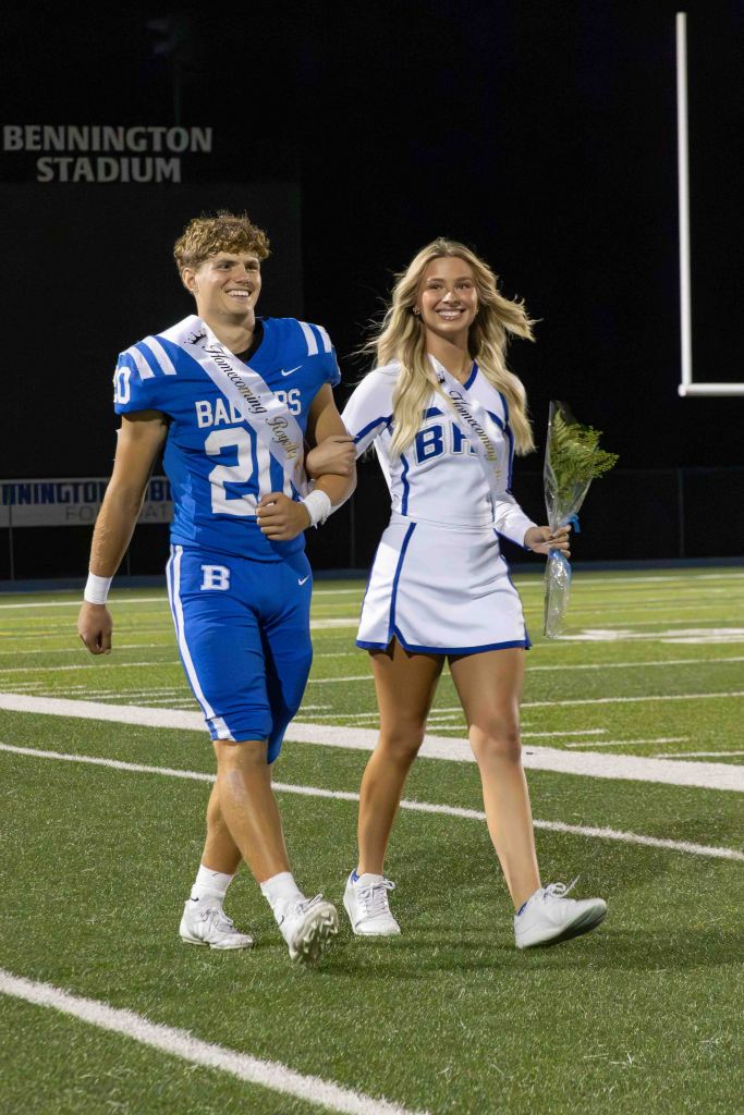 A male high school football player in a blue uniform with the number 20, wearing a sash, walks alongside a female cheerleader in a white outfit, also wearing a sash, both smiling on a football field at night.
