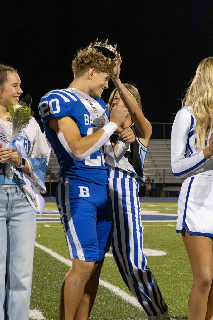 A football player in a blue jersey receiving a crown during a ceremony, smiling at a girl who is placing the crown on his head, with other students and cheerleaders nearby.