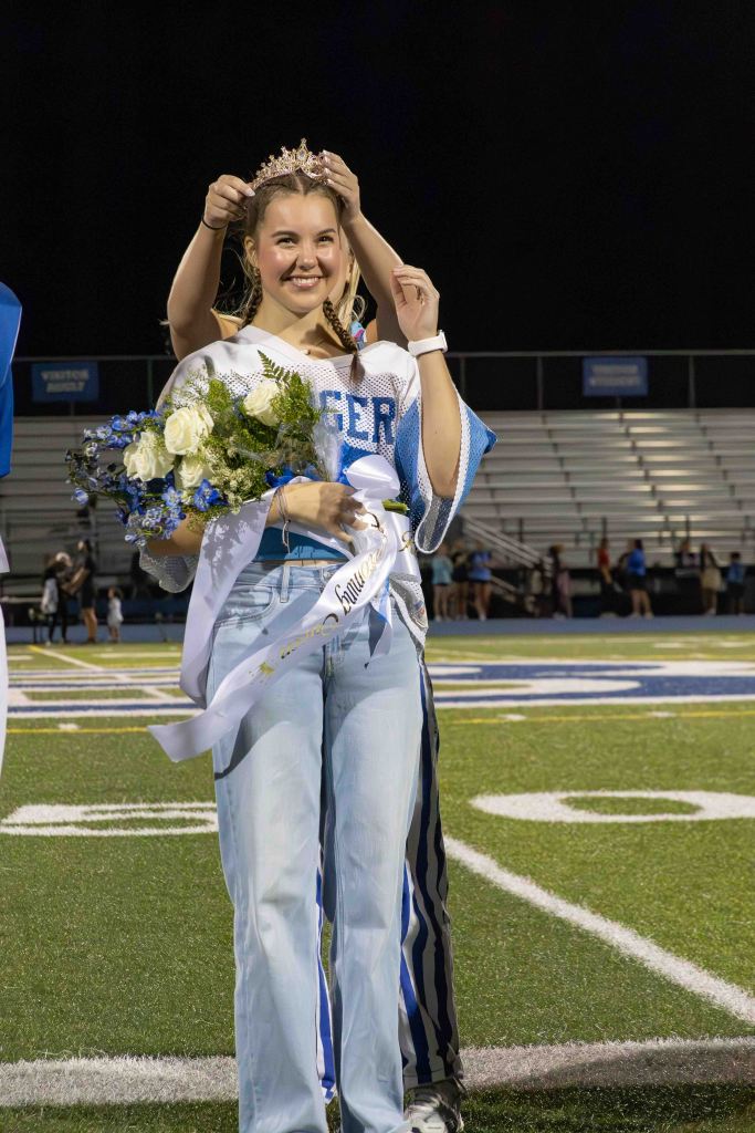 A young woman wearing a tiara and holding a bouquet of flowers smiles as she is crowned at a football field during a night event.