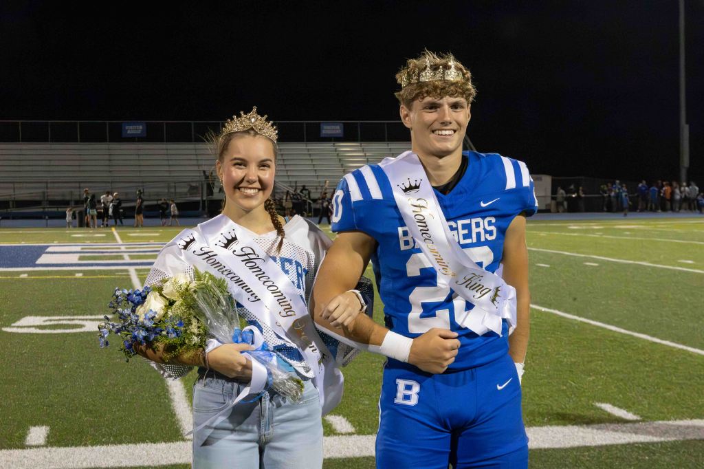 A girl crowned as Homecoming Queen wearing a sash and holding flowers, stands beside a boy crowned as Homecoming King in a football uniform, both smiling on a football field at night.