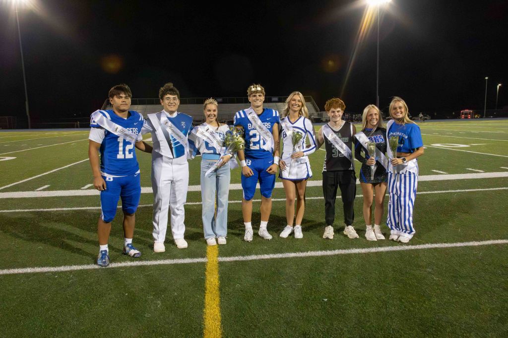 Group of students in athletic and cheerleading uniforms posing on a football field during a night event, some holding flowers and wearing sashes.