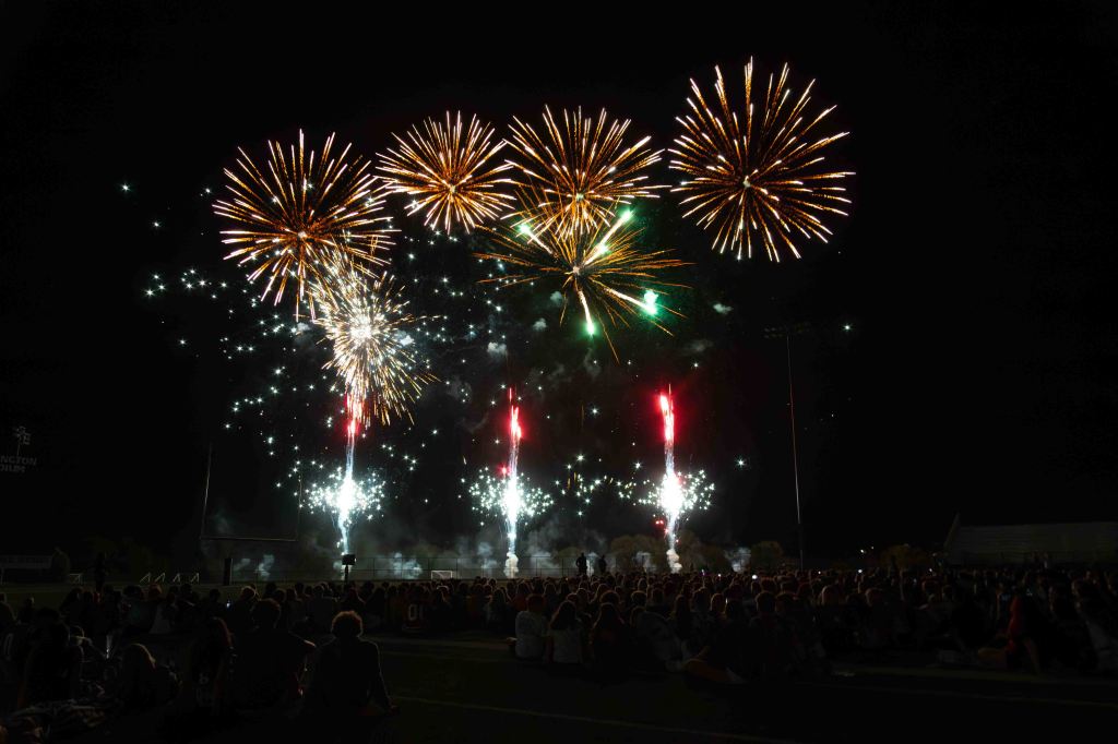 A vibrant fireworks display lighting up the night sky above a large crowd of spectators sitting on a field.