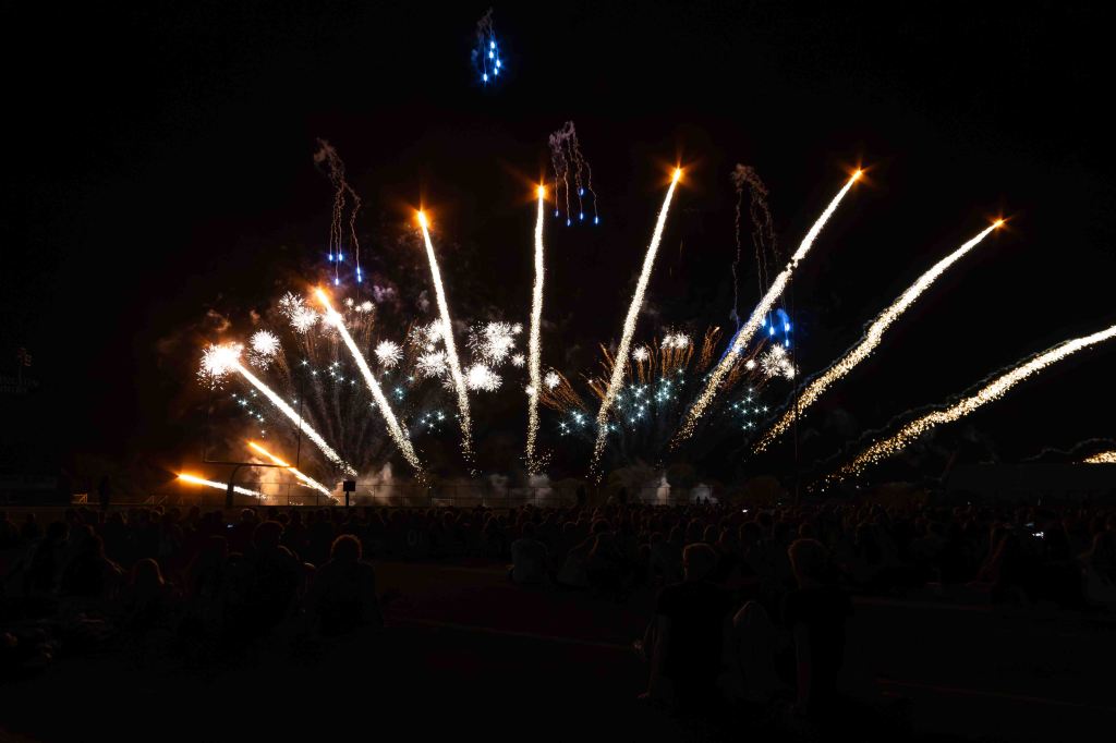A vibrant fireworks display lighting up the night sky, with colorful sparks and trails against a dark background, while a large audience watches from below.