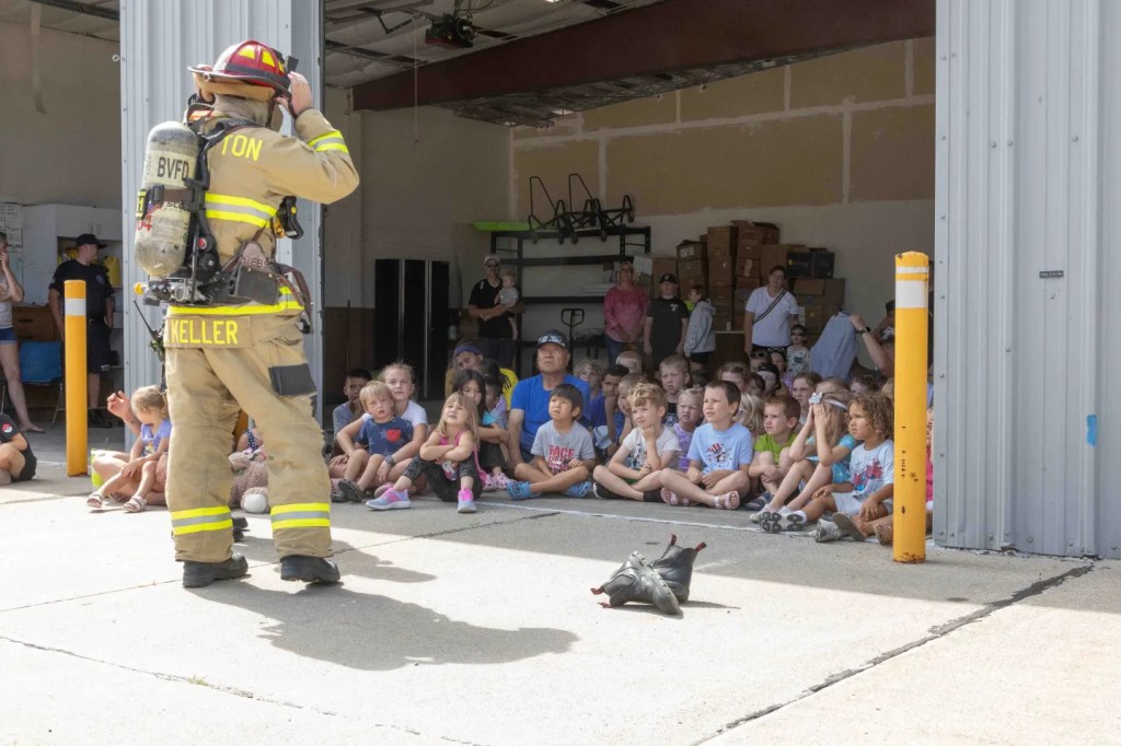 A firefighter in full gear addresses a group of seated children in a garage-type facility, with onlookers in the background.