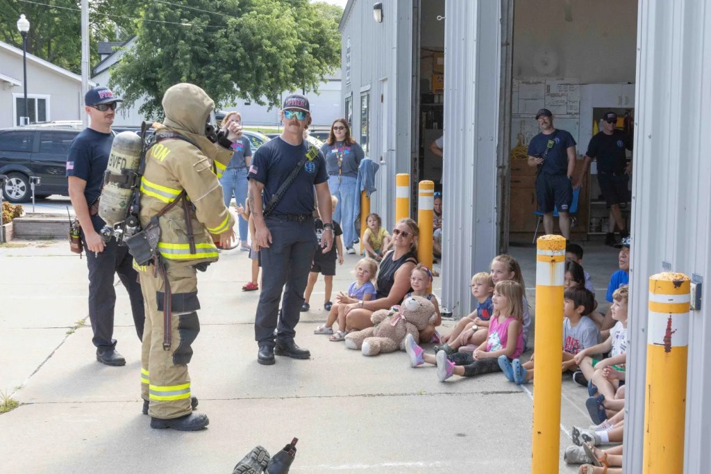 A group of young children sitting on the ground, watching a firefighter in full gear, while adults and other firefighters are present, outside a fire station.