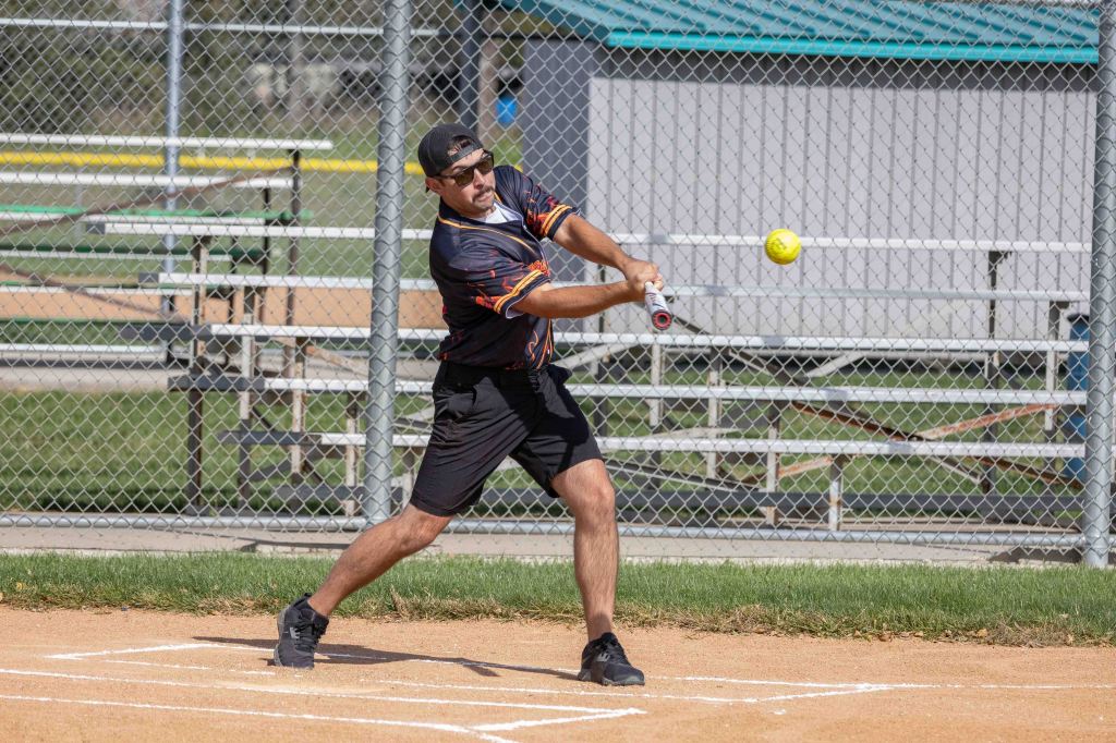 A man in a black and orange sports jersey swinging a bat at a yellow softball in a baseball diamond, with bleachers in the background.