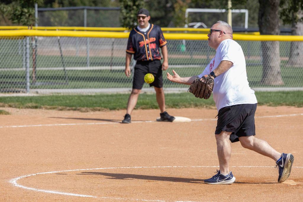 A man in a white t-shirt and black shorts is pitching a softball on a dirt field, while another man in a black jersey with orange accents stands ready in the background.