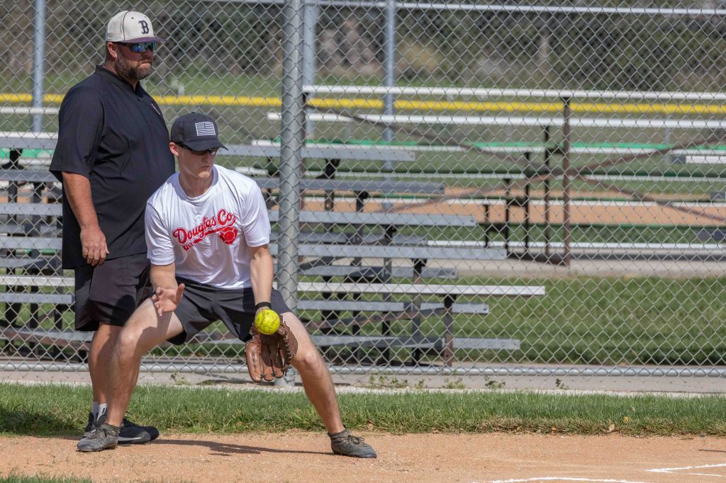 A coach stands behind a young player in a white shirt with red lettering, preparing to field a softball. The player is in a crouched position on a baseball field, with empty bleachers visible in the background.