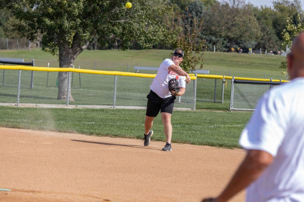 A player wearing a white shirt and black shorts is throwing a softball from the infield, with a fence and trees in the background.