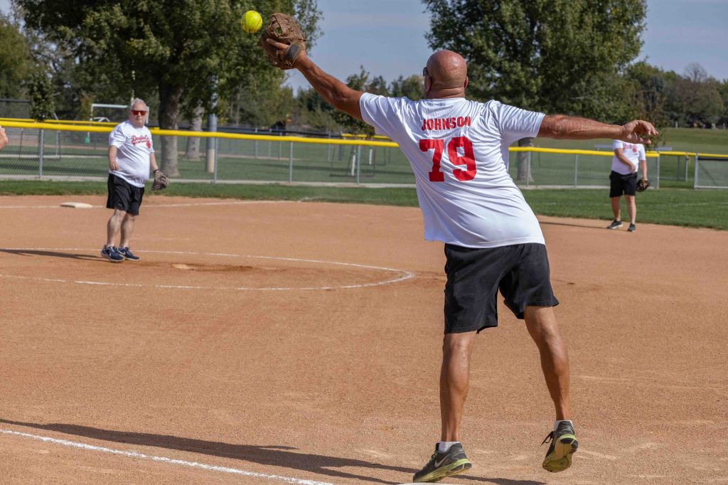 A player wearing a white shirt with the name 'Johnson' and the number '79' throws a yellow softball while standing on a sandy field, with other players in the background.