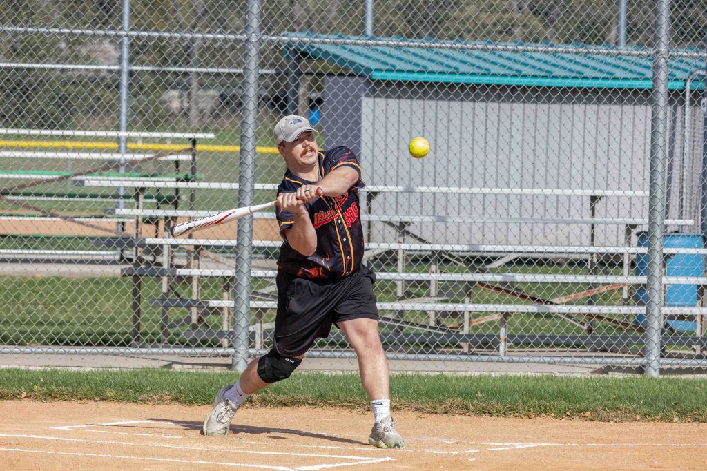 A player swings a bat to hit a softball on a baseball field with bleachers in the background.