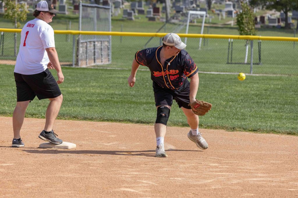 A softball player in a black jersey throws a yellow ball while another player in a white shirt stands on base at a softball field.