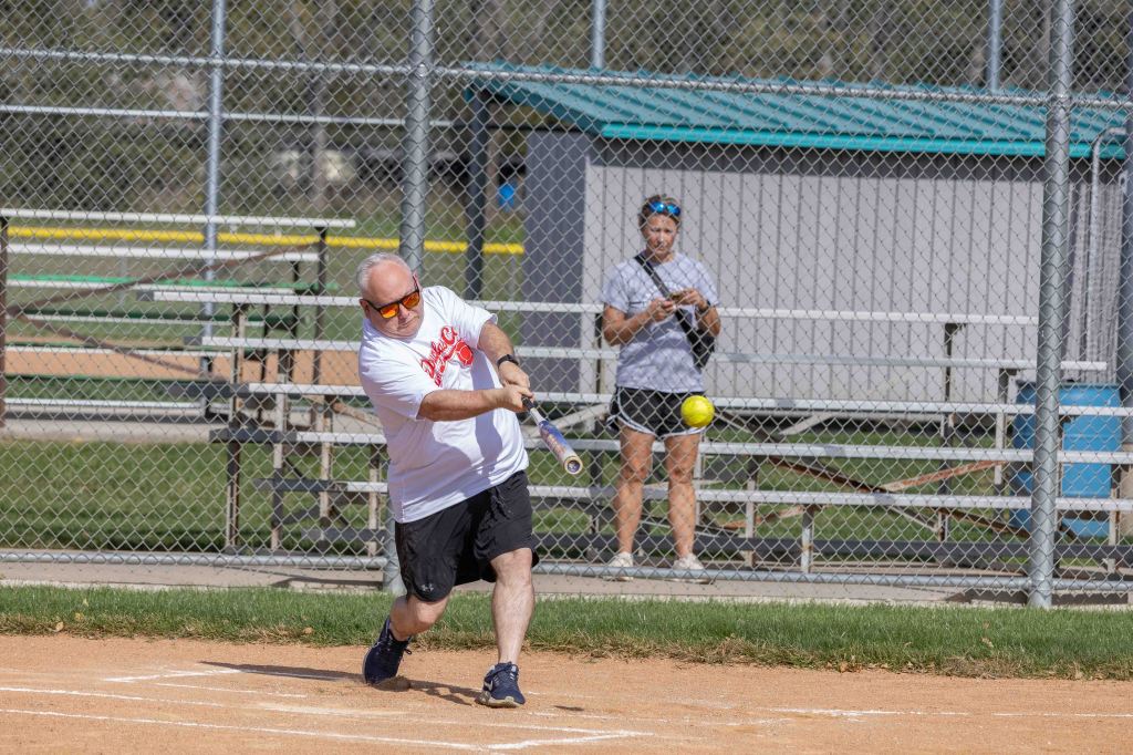 A man swings a bat, hitting a softball in a sports field while a woman watches from the background.