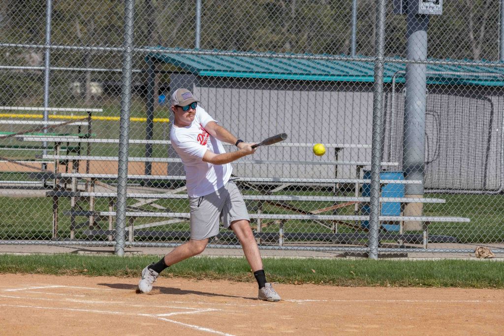 A young man in a white shirt and gray shorts swings a baseball bat at a yellow softball on an outdoor field surrounded by a chain-link fence.