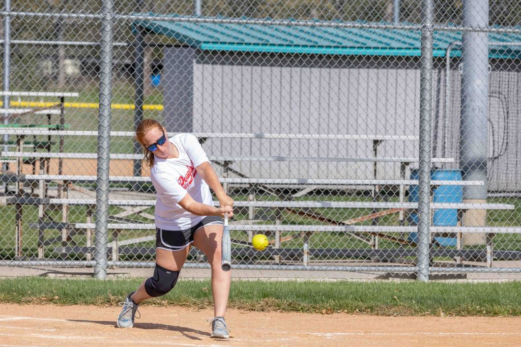 A female softball player in a white t-shirt swings a bat at a yellow softball on a dirt field, with bleachers and a shelter in the background.