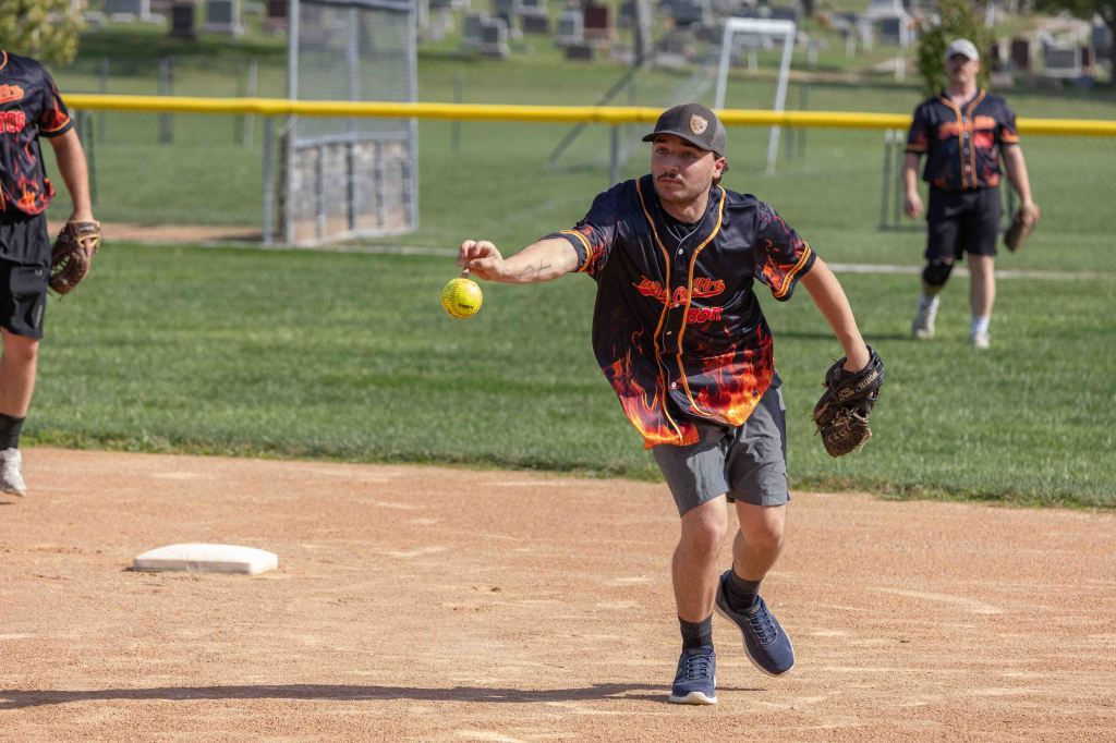 A player in a fiery-themed jersey throws a softball while standing on an infield diamond during a game.