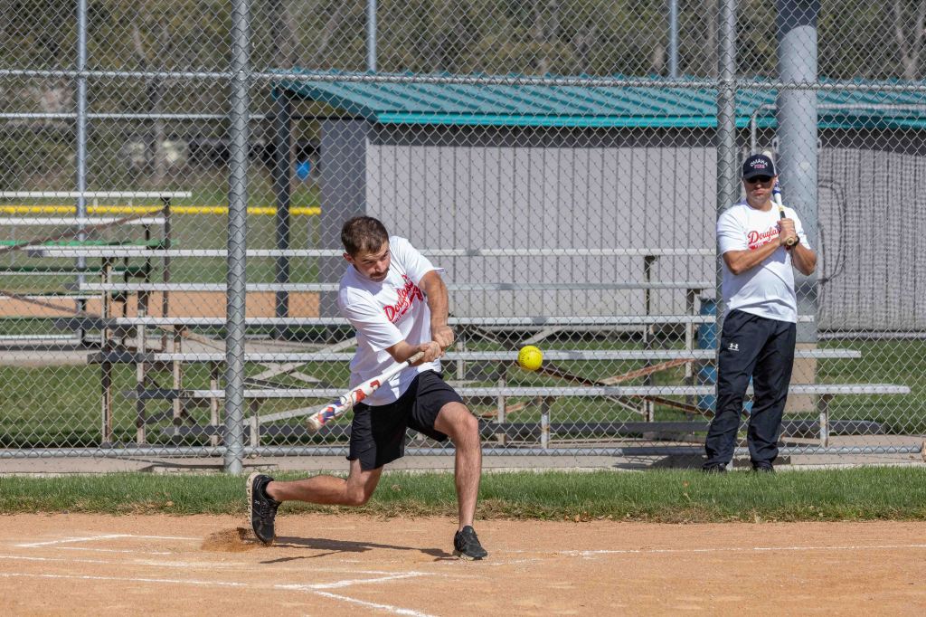 A young man swinging a bat to hit a softball at a baseball field, while another person stands nearby watching.