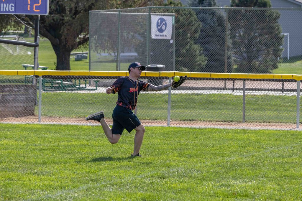 A player in a baseball uniform is running and catching a softball near the outfield fence, with a scoreboard in the background displaying the inning.