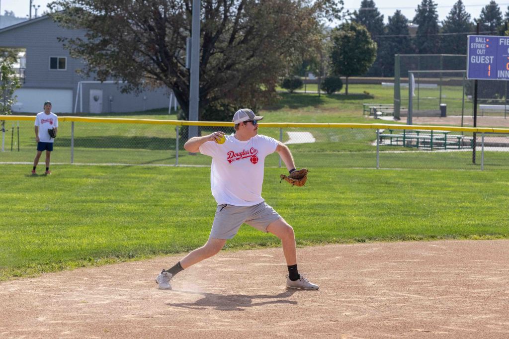 A young male baseball player in a white shirt and grey shorts is throwing a baseball on a field. Another player is seen in the background. The scene is set on a sunny day with a grassy area and a baseball diamond.