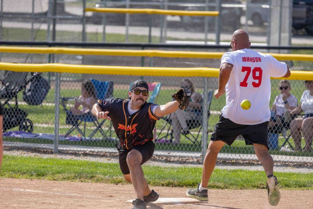 A baseball player in a black jersey reaches for a thrown ball while another player in a white jersey prepares to run towards first base, with spectators in the background.