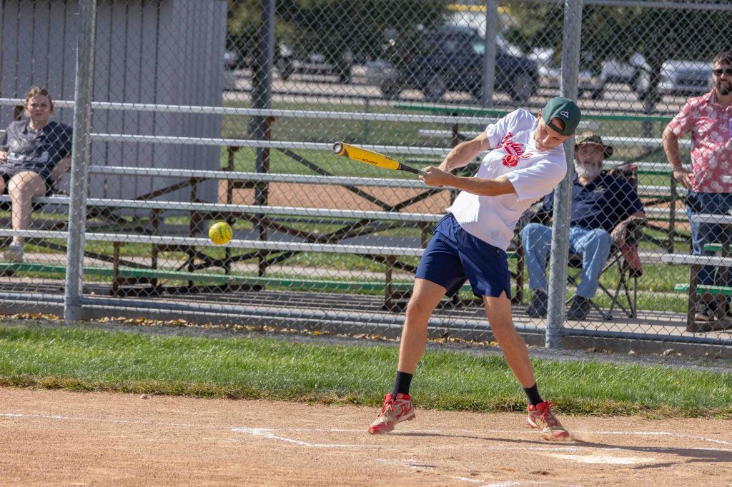 A young man swings a bat at a yellow softball during a game, with spectators seated in the background.