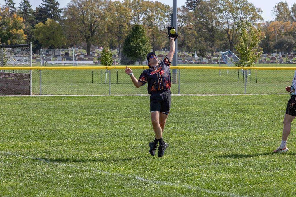 A player in a black and orange softball uniform jumps to catch a flying softball in a grassy field, with trees and a cemetery in the background.