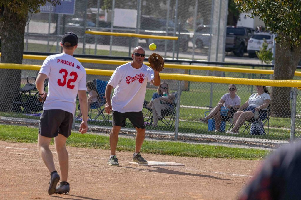 A man in a white t-shirt catching a softball while another player in a red jersey runs towards him at a softball field.