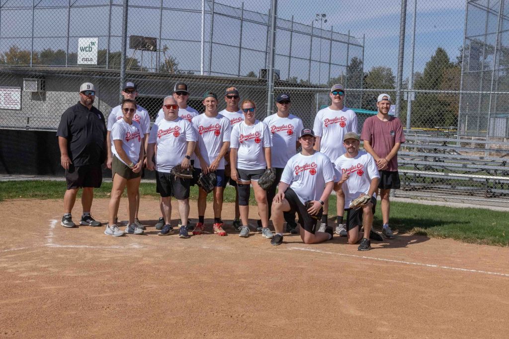 A group of ten individuals poses on a baseball field, wearing matching white t-shirts with 'Douglas Co' printed on them. They are smiling and some hold baseball gloves. The scene is set against a backdrop of a baseball diamond and bleachers.