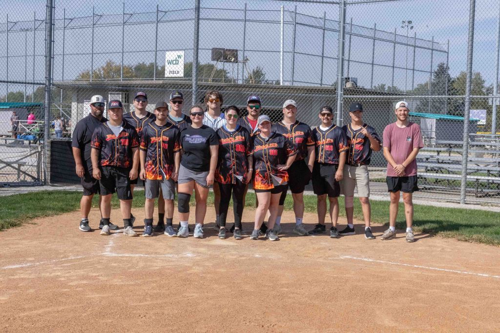 A group of eleven people posing on a softball field, wearing matching jerseys with a flame design, smiling and standing together in front of a chain-link fence.