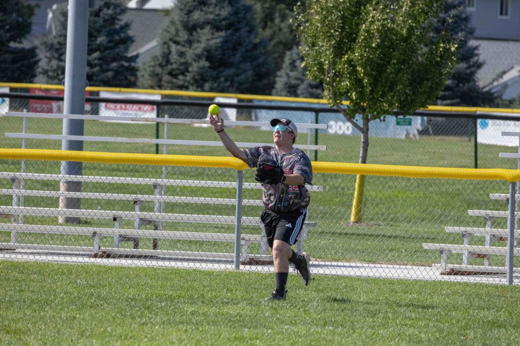 A young player in a baseball uniform reaches up to catch a yellow softball while playing in an outdoor field with bleachers in the background.
