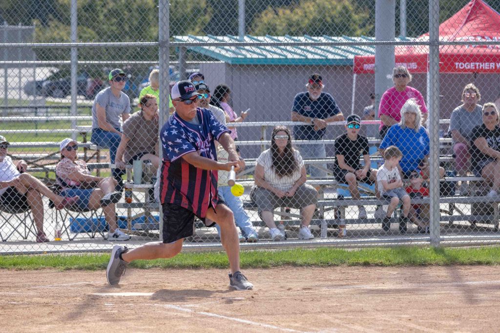 A player in an American flag-themed shirt swings a bat at a softball during a game, with a crowd of spectators seated in bleachers behind him.