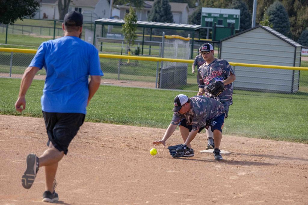 Three players participating in a softball game on a dirt field, with one player diving to catch a yellow ball while another runs in the background.
