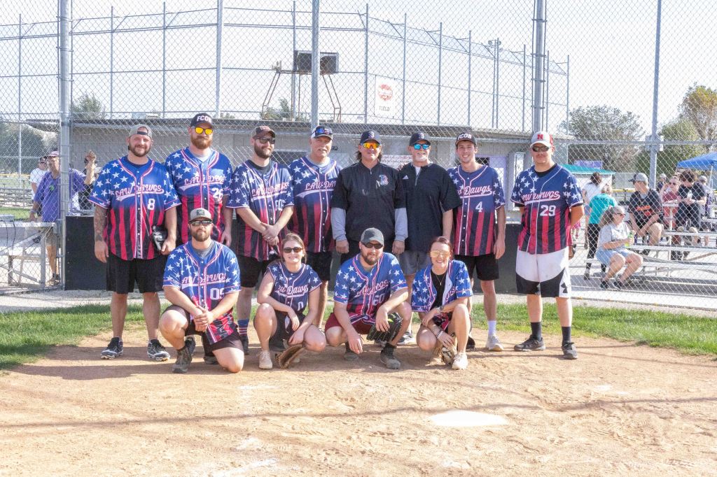 A group of softball players standing together on a field, wearing matching jerseys with red, white, and blue colors. The team poses on a dirt area near home plate, with spectators and a baseball field visible in the background.