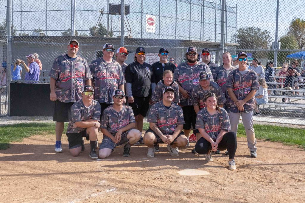A group photo of a softball team, featuring players in matching camo jerseys and caps. They are posing on a baseball field with a fence and spectators in the background.