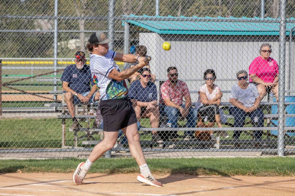A player striking a softball in a game while spectators watch from behind a chain-link fence.