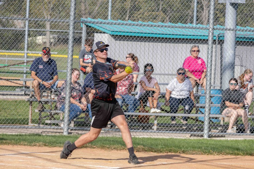 A softball player in a black jersey swings at a pitch on a field, with spectators watching from the bleachers in the background.