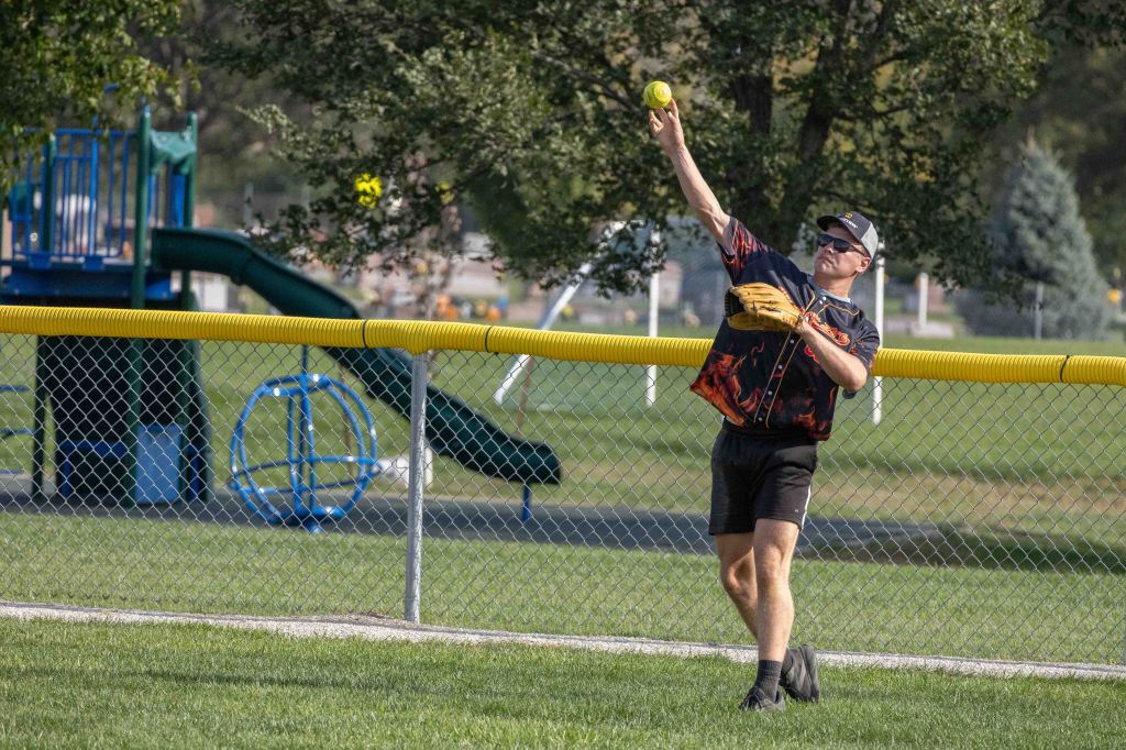 A player in a black and red patterned shirt and sunglasses throws a softball while standing near a chain-link fence at a park, with a playground structure in the background.