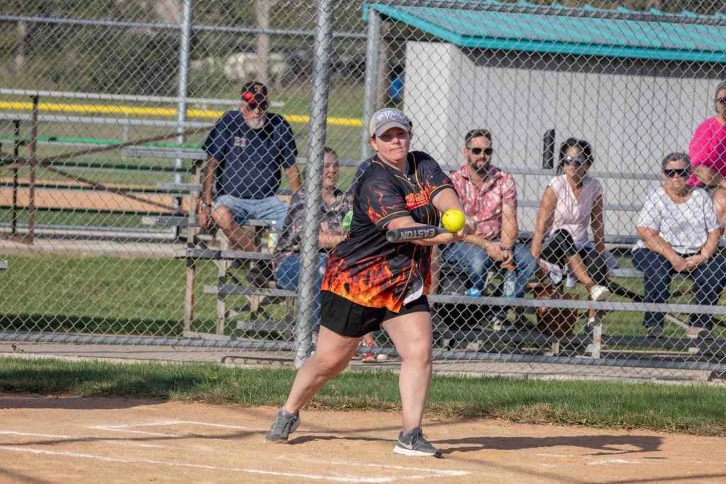 A woman in a black and orange softball jersey swings a bat at a yellow softball on a dirt field, with spectators sitting in the background.