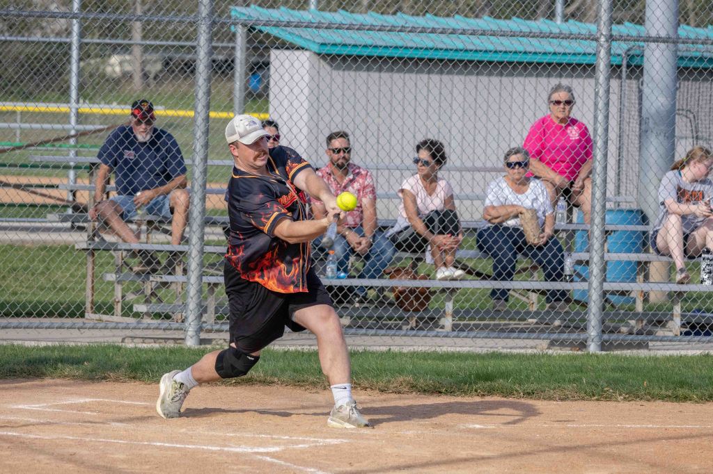 A player in a black and orange jersey swings a bat at a yellow ball on a softball field, with spectators seated in the background.