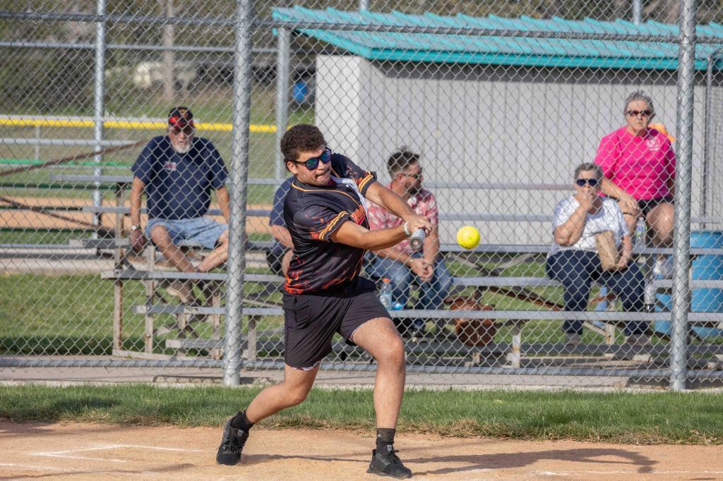 A young man swings a bat at a softball on a diamond, with spectators watching from behind a fence.