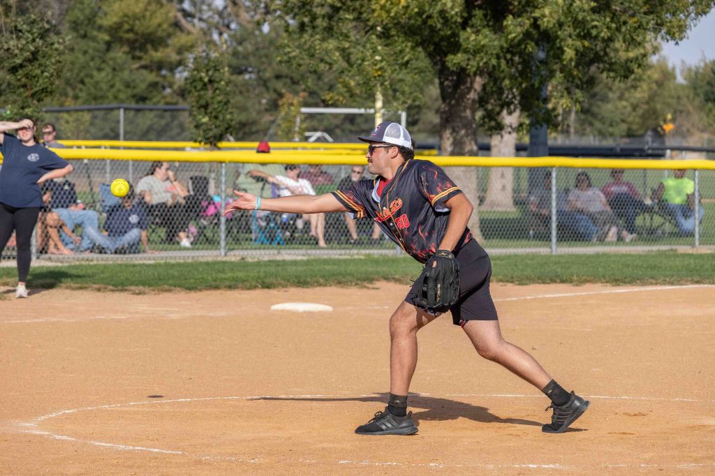 A male softball player wearing a black jersey and shorts is pitching a yellow softball on a dirt field, with spectators in the background.