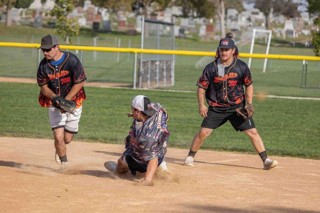 Three male softball players in action during a game, one sliding on the dirt, while the others field the ball. The scene is set on a sunny day with a grassy field and a cemetery visible in the background.