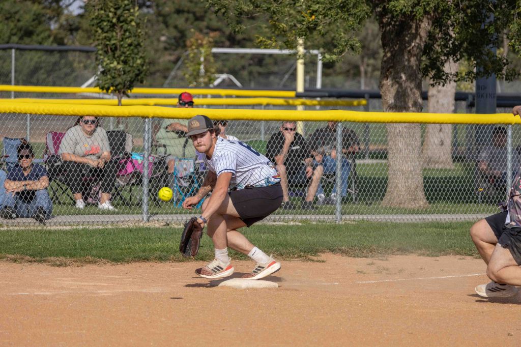 A player crouches on the baseball field, preparing to catch a yellow softball while spectators watch from behind a fence.