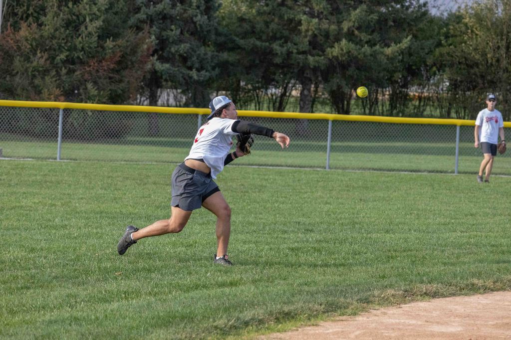 A player in a black long-sleeve shirt and gray shorts is throwing a yellow softball while running on a grass field. Another player is in the background, preparing to catch the ball.