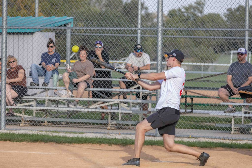 A young man in a white shirt and black shorts swings a bat at a yellow softball, while spectators watch from the bleachers in a softball field.