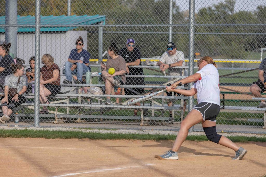 A female softball player swinging a bat to hit a ball during a game, with a crowd of spectators sitting on bleachers in the background.