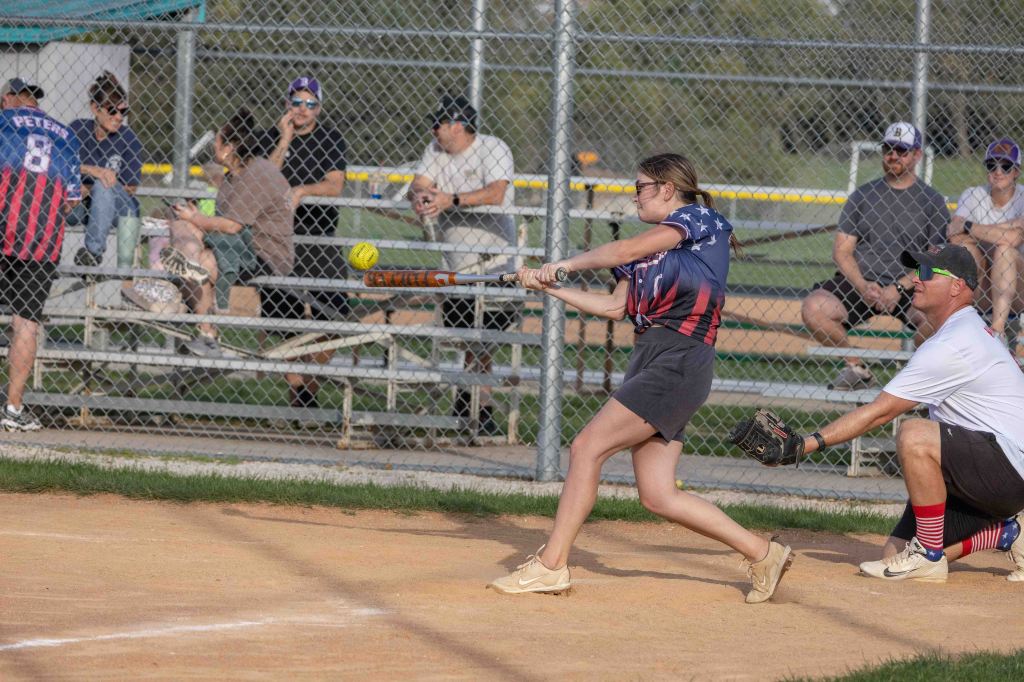 A woman wearing an American flag-themed jersey swings a bat at a softball while a player crouches in a catching position, with seated spectators in the background.